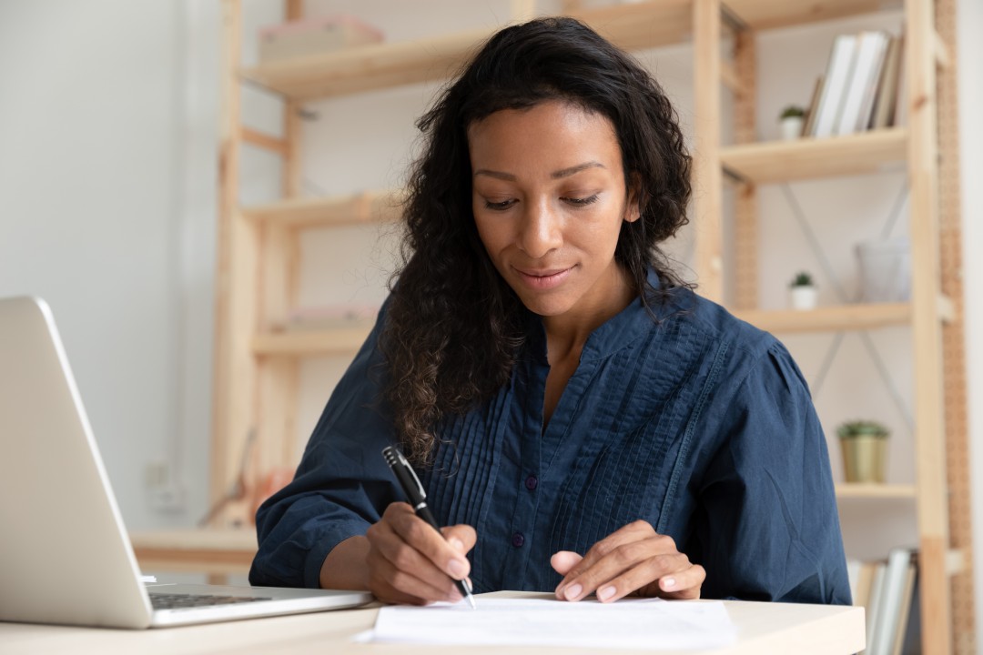 Woman writing notes at her desk with laptop open