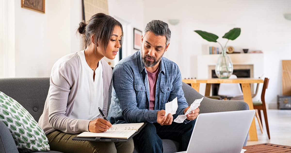 A man and a woman sit on a couch in front of a laptop computer looking through receipts and writing in a notebook.