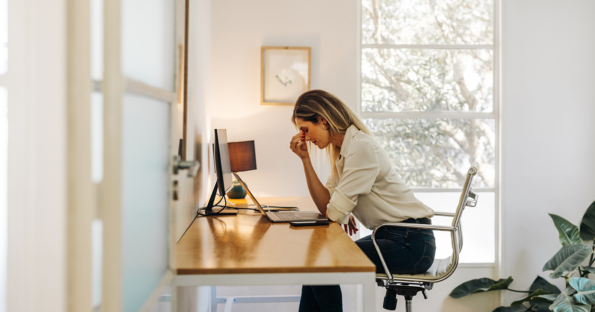 A woman sitting at a desk leans forward and, using her right hand, squeezes the area where her nose meets her forehead.