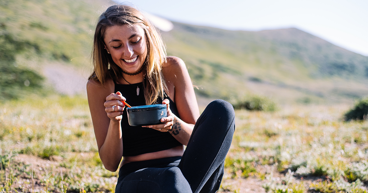 A woman sits outside on the ground happily eating out of a bowl.