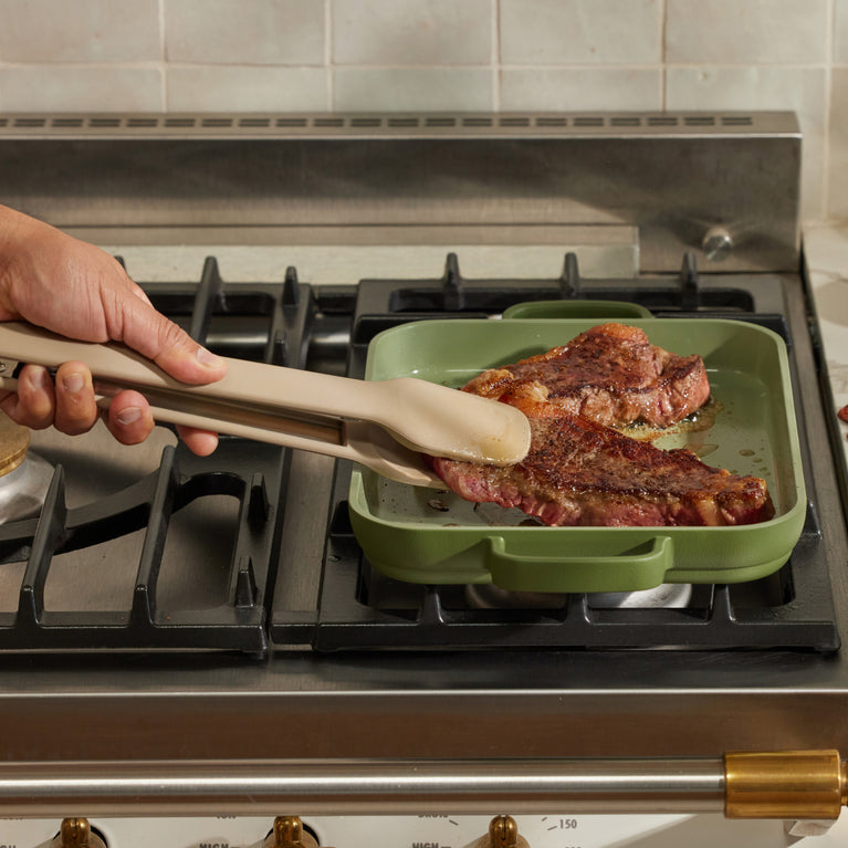 beige tongs flipping steak on green griddle pan on stovetop