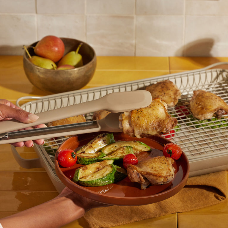 beige tongs picking up cooked chicken from beige griddle pan and oven rack on counter