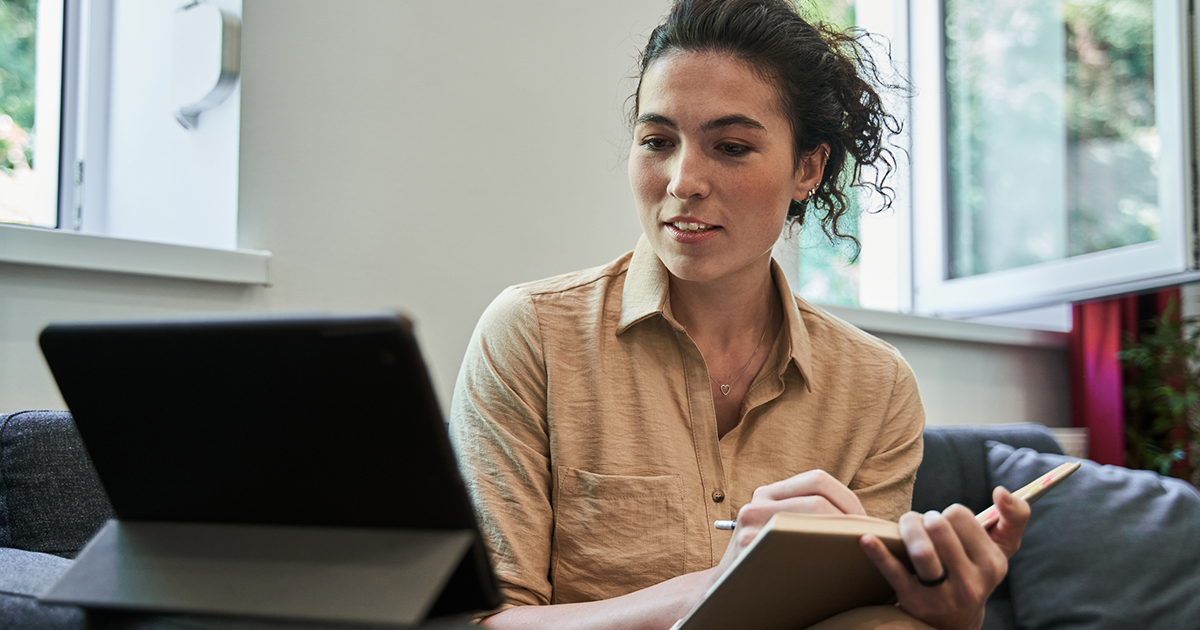 A woman video chats with someone on a table while taking notes in a notebook.