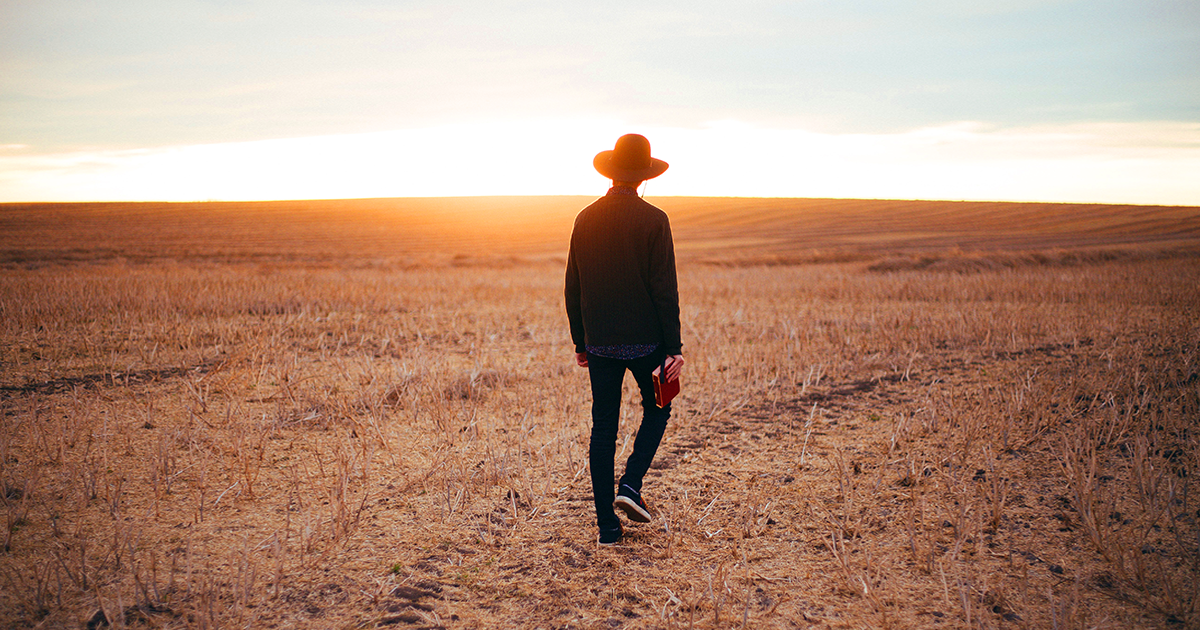 A man in a wide-brim hat walks in a field holding a book in his right hand.