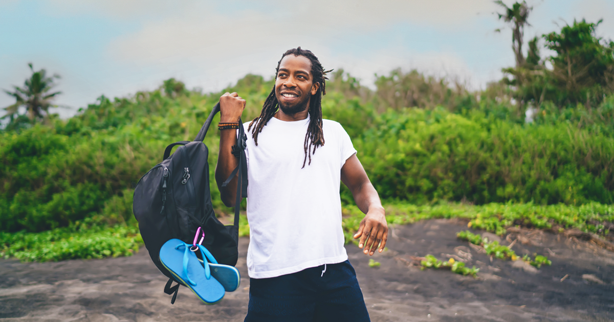 A man outside in nature lifts a black backpack onto one shoulder. Blue flip-flops are attached to the bag by a purple carabiner.